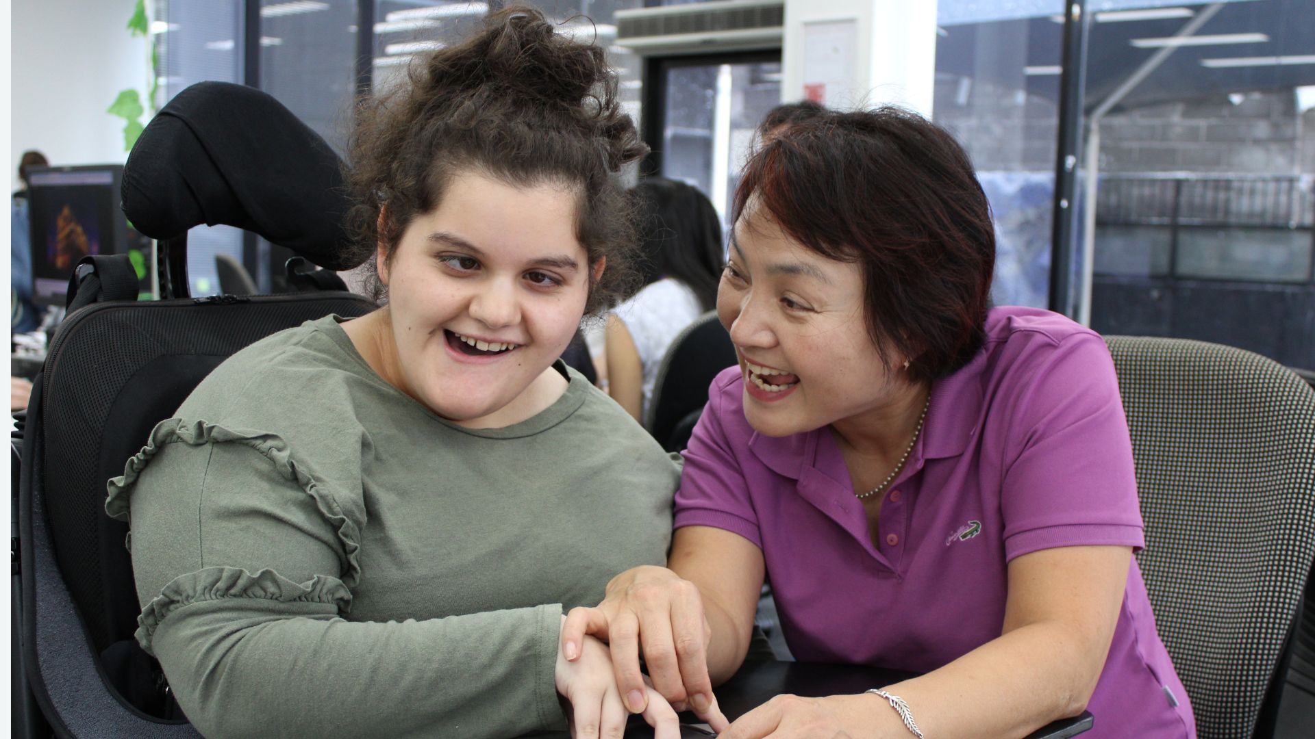 A lady with wavy dark hair and a khaki long sleeved tshirt is in a wheelchair smiling at the camera. Her support worker is holding her hand smiling at her; she has short black hair and is wearing a purple t shirt.